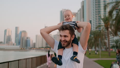 Glückliche-Junge-Familie-Liebenden-Jungen-Vater-In-Weißem-T-Shirt-Und-Borsten-Hipster-Zu-Fuß-Mit-Seinem-Sohn-Sitzt-Auf-Seinem-Hals-An-Der-Uferpromenade-Im-Hintergrund-Der-Modernen-Stadt-Im-Sommer-An-Der-Uferpromenade