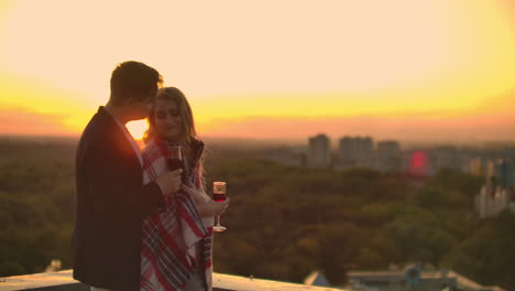 Man-and-young-pretty-woman-sitting-on-couch-holding-wine-glasses-and-kissing-on-rooftop-terrace-at-sunset