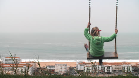 Little-girl-on-swing-overlooking-Nazare-ocean-coast-with-hotels-Portugal