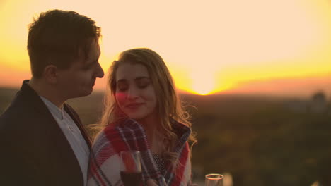 A-young-couple-on-the-roof-drinking-wine-from-glasses-standing-in-a-blanket-and-admiring-the-beautiful-sunset-over-the-city.-Romantic-evening-on-the-roof-overlooking-the-city