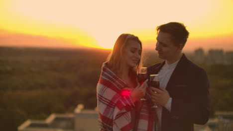 A-young-couple-on-the-roof-drinking-wine-from-glasses-standing-in-a-blanket-and-admiring-the-beautiful-sunset-over-the-city.-Romantic-evening-on-the-roof-overlooking-the-city