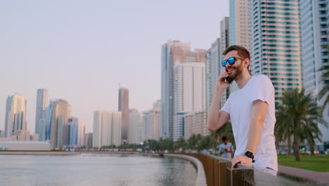 Joven-Turista-Con-Gafas-De-Sol-Y-Cerdas-Hipster-Con-Camiseta-Blanca-Parado-En-El-Paseo-Marítimo-En-El-Fondo-De-La-Ciudad-Moderna-Hablando-Por-Teléfono-Y-Escuchando-La-Respuesta