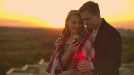 Una-Mujer-Con-Copas-De-Vino-Está-En-El-Techo-Y-Un-Hombre-Le-Pone-Una-Manta-Sobre-Los-Hombros-Y-Lo-Abraza.-Una-Pareja-Amorosa-Bebe-Vino-Y-Se-Abraza-Al-Atardecer-En-El-Techo