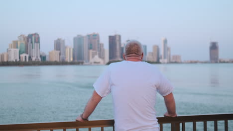 Älterer-Mann-Im-Weißen-T-Shirt-Mit-Blick-Auf-Die-Bucht,-Der-Am-Wasser-Steht-Und-Die-Rückansicht-Mit-Blick-Auf-Die-Schöne-Aussicht-Betrachtet