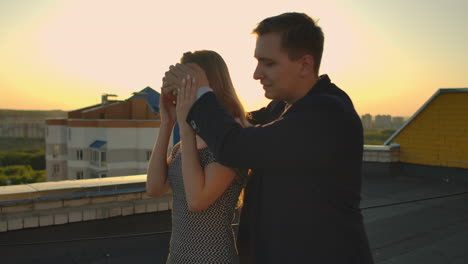 A-date-on-the-roof-a-man-is-leading-girl-closing-hands-eyes-preparing-romantic-surprise.-Show-a-beautiful-view-at-sunset.-The-woman-enjoys-the-surprise-and-smiles-looking-at-the-beautiful-view-and-the-man.