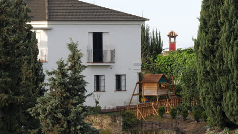 Exterior-De-Una-Casa-De-Dos-Plantas-Con-Espacio-Para-Niños-Y-Grandes-Pinos-A-Un-Lado.