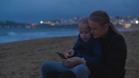 Mother-and-daughter-watching-something-on-smartphone-at-ocean-beach