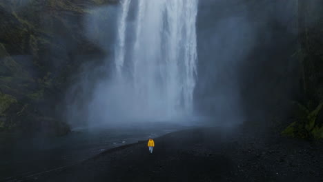 Un-Hombre-Con-Chaqueta-Amarilla-Camina-Cerca-De-Una-Cascada-Sobre-Espectaculares-Acantilados-De-Roca-Negra-En-Skógafoss,-Islandia