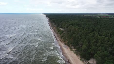 Revealing-a-majestic-cliff-on-the-Baltic-Sea-coastline-in-Lithuania-through-drone-footage,-the-scene-unfolds-with-tumultuous-waves-and-the-vivid-green-tones-of-the-water-under-moody-skies