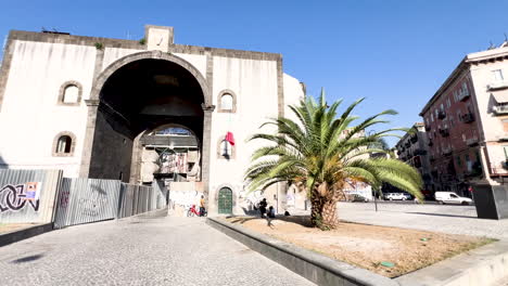 Porta-Capuana-in-Naples,-an-ancient-city-gate,-showcases-the-rich-history-and-architecture-under-a-bright-blue-sky,-with-a-striking-palm-tree-adding-to-the-scene's-Mediterranean-charm