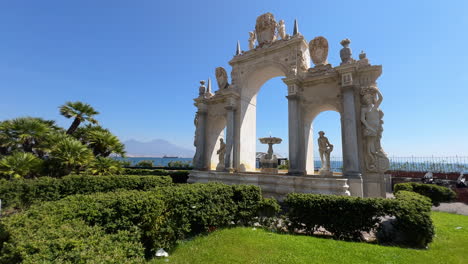Elegante-Arco-Del-Sebeto-Y-Estatuas-En-Un-Jardín-Iluminado-Por-El-Sol-Con-Una-Vista-Impresionante-Del-Mar-Y-El-Monte-Vesubio-En-La-Distancia