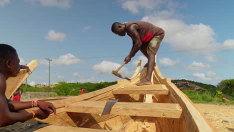 Black-african-man-carving-wooden-boat-with-adze,-another-man-watching