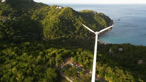 Aerial-orbiting-shot-of-wind-mill-on-koh-tao-island-with-bay-and-green-mountains-in-background-at-sunset-time,-Thailand