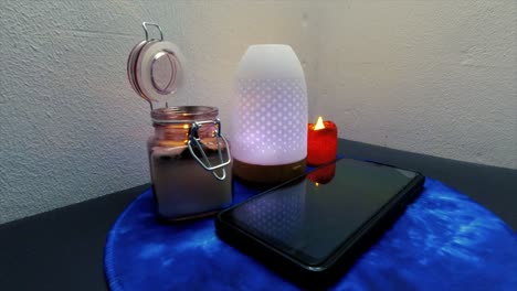 black-mobile-cellphone-that-lights-up-incoming-caller-surrounded-by-lights-and-humidifier-that-changes-in-colors-and-an-open-lit-candle-in-a-glass-jar