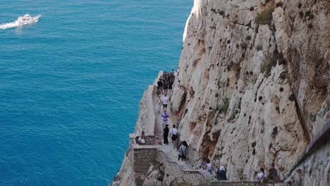 Many-tourists-walking-on-a-path-between-a-rock-wall-and-the-blue-sea