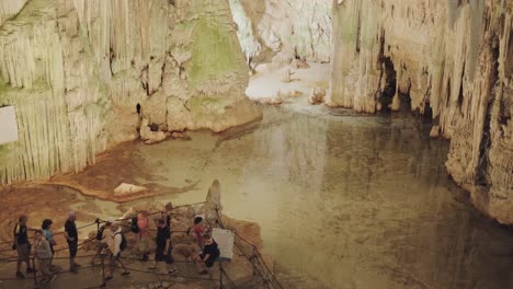 Varios-Turistas-Caminan-Con-Un-Guía-Turístico-Por-La-Gruta-De-Neptuno,-Con-Estalactitas-Y-Junto-A-Una-Laguna-Subterránea.