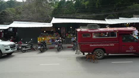 Red-van-driving-past-local-shops-and-motorbikes-on-a-busy-street-in-the-city-of-Chiang-Mai,-Thailand