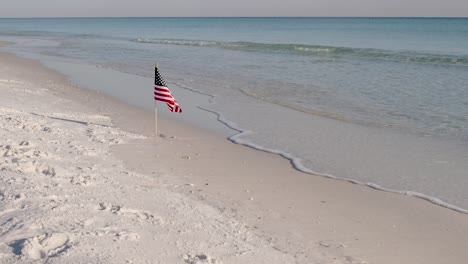 Fly-over-the-American-flag-on-a-white-sand-beach-with-clear-waters