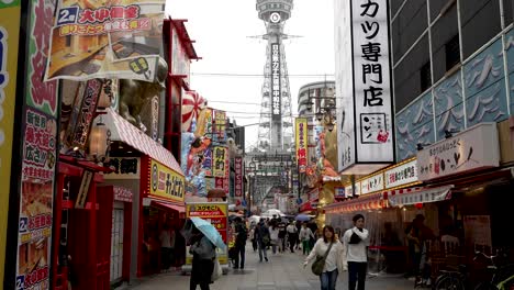 Besucher-Erkunden-Shinsekai-An-Einem-Bewölkten,-Regnerischen-Tag-Mit-Blick-Auf-Den-Tsutenkaku-Turm-Im-Hintergrund