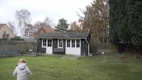 Toddler-walking-in-the-garden-with-an-old-garden-house-in-the-background,-storage-room-for-gardening-tools