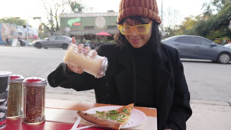 Cute-Asian-woman-shakes-parmesan-topping-onto-margherita-pizza-slice-at-outdoor-street-pizza-restaurant