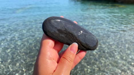 Hand-holding-and-playing-with-a-wet-smooth-black-rock-at-the-beach-with-turquoise-sea-water-in-Manilva-Spain,-sunny-summer-day,-4K-static-shot