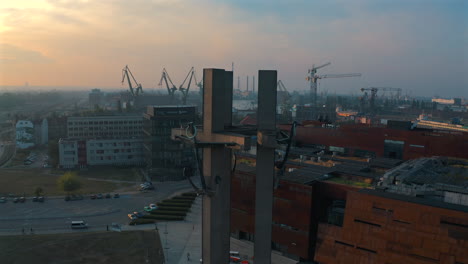 Aerial-shot-of-crosses-near-Museum-of-Solidarity-in-Gdańsk,-Poland-with-shipyard-and-cranes-in-the-background