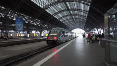 Tren-Eléctrico-Que-Sale-De-La-Pintoresca-Estación-De-Tren-De-Leipzig-Con-Viajeros-Caminando-Sobre-El-Andén-En-Un-Día-Brillante,-Europa,-Alemania