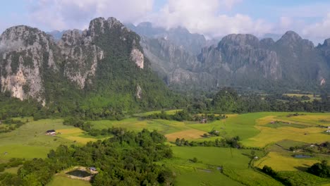 Aerial-View-Of-Green-Farming-Fields-With-Soaring-Forested-Mountain-Landscape-At-Vang-Vieng
