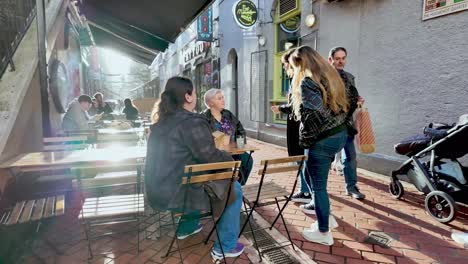 Frühe-Morgensonne-Auf-Der-Straße-In-Cork-City-Und-Eine-Gruppe-Junger-Frauen,-Die-Auf-Der-Terrasse-Eines-Pubs-Sitzen,-Während-Fußgänger-In-Zeitlupe-Vorbeikommen