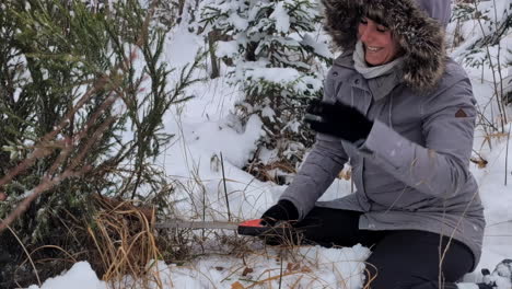 Glückliche,-Lächelnde-Frau,-Die-In-Einem-Verschneiten-Wald-Mit-Einer-Säge-Einen-Weihnachtsbaum-Fällt