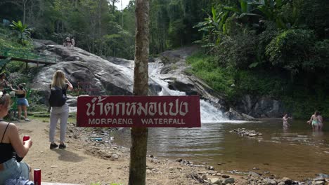 Vista-De-Un-Turista-Parado-Junto-Al-Cartel-Con-El-Nombre-De-La-Cascada-Mo-Pang,-Con-Un-Bosque-Verde-Al-Fondo