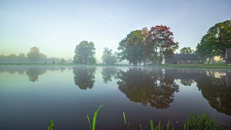 Time-lapse-of-a-misty-sunrise-at-a-mirroring-lake,-fall-colors-at-rural-house