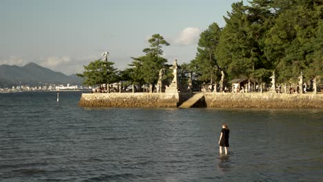 Turista-Femenina-Caminando-Hacia-Las-Aguas-De-La-Marea-Entrante-En-El-Santuario-Itsukushima-Durante-La-Hora-Dorada