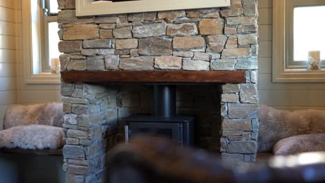 Slow-reveal-of-a-new-black-rustic-log-burner-fire-in-a-hand-built-stone-wall-fire-place-of-old-fashioned-farm-house-surrounded-by-chairs,-logs-and-pillows