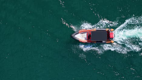 Inflatable-orange-red-black-coastguard-boat-drives-across-glistening-ocean,-drone-top-down-static