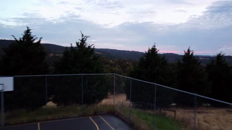 Luftaufnahme-Eines-Basketballplatzes-Mit-Einem-Schönen-Blick-Auf-Die-Bäume-Und-Hügel-Dahinter-Mit-Einem-Hauch-Des-Rosafarbenen-Sonnenuntergangs