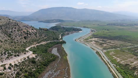 Drone-view-in-Albania-flying-over-wide-blue-river-and-green-landscape-next-to-the-sea-and-road-with-mountains-on-the-back