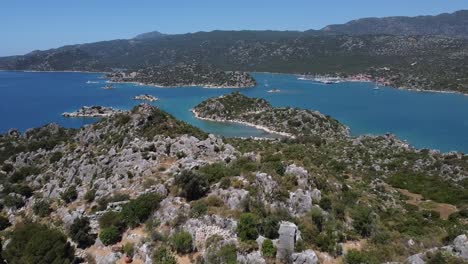 Aerial-tour-over-Aperlai-Ancient-City-with-mountains,-blue-waters-and-boats-sailing-in-Turkey
