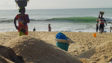 African-woman-carrying-basket-of-sand-on-head,-emptying-on-beach-pile