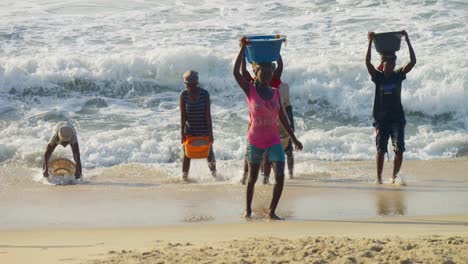 Mujeres-Africanas-Cargando-Cestas-De-Arena-En-La-Cabeza,-Saliendo-De-Las-Olas-Del-Mar