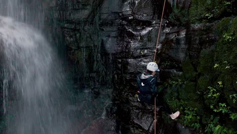 Vista-Aérea-De-Una-Escaladora-Bajando-Por-La-Pared-De-La-Montaña-Junto-A-La-Cascada