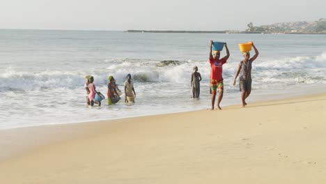 Mujeres-Africanas-Cargando-Cestas-En-La-Cabeza-En-La-Playa,-Otras-Esperan-En-Olas