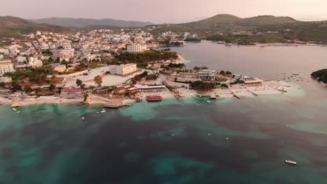 Drone-view-in-Albania-flying-over-blue-crystal-clear-water-on-sunset,-green-hills,-boats-and-hotels-in-Ksamil