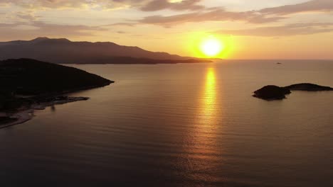 Drone-view-in-Albania-flying-over-blue-crystal-clear-water-on-sunset,-with-the-sun-setting-in-the-horizon-with-an-island-in-Ksamil