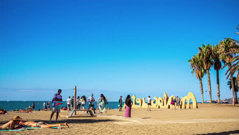 Gente-De-Lapso-De-Tiempo-Moviéndose-En-La-Playa-Cerca-Del-Cartel-De-Málaga-En-España,-Mar-Mediterráneo