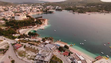 Drone-view-in-Albania-flying-over-blue-crystal-clear-water-on-sunset,-green-hills,-boats-and-hotels-in-Ksamil