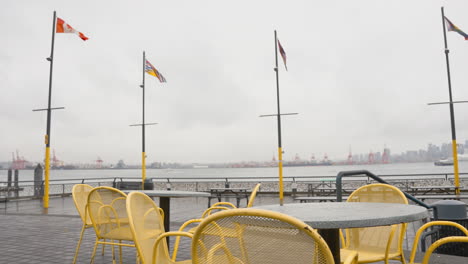 Una-Toma-De-Izquierda-A-Derecha-De-Sillas-Amarillas-Vacías-En-Un-Muelle-Contra-El-Horizonte-De-La-Ciudad-De-Vancouver,-Con-El-Seabus-Y-Varias-Banderas-Visibles