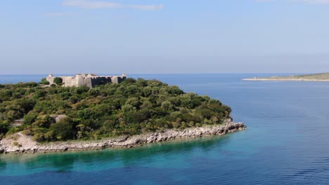 Drone-view-in-Albania-flying-over-blue-clear-ocean-with-a-small-green-island-with-a-medieval-fort-on-it-on-a-sunny-day