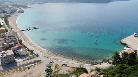 Drohnenansicht-In-Albanien,-Die-An-Einem-Sonnigen-Tag-über-Einen-Strand-Mit-Kristallklarem,-Blauem-Wasser,-Gebäuden-Am-Hafen-Und-Einer-Grünen-Insel-Fliegt